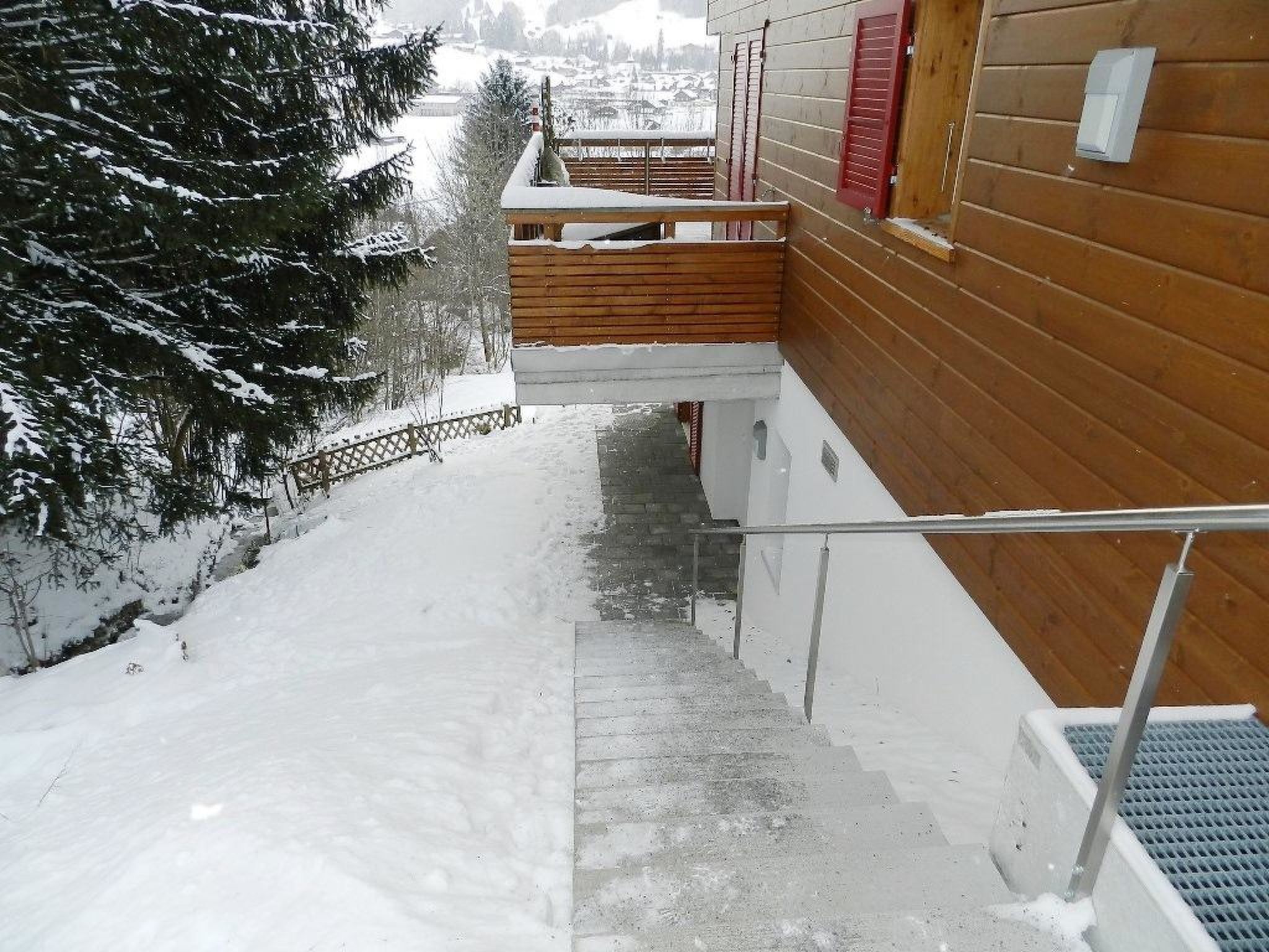 A wooden staircase leading to a wooden deck with a metal railing, surrounded by snow-covered ground and trees.