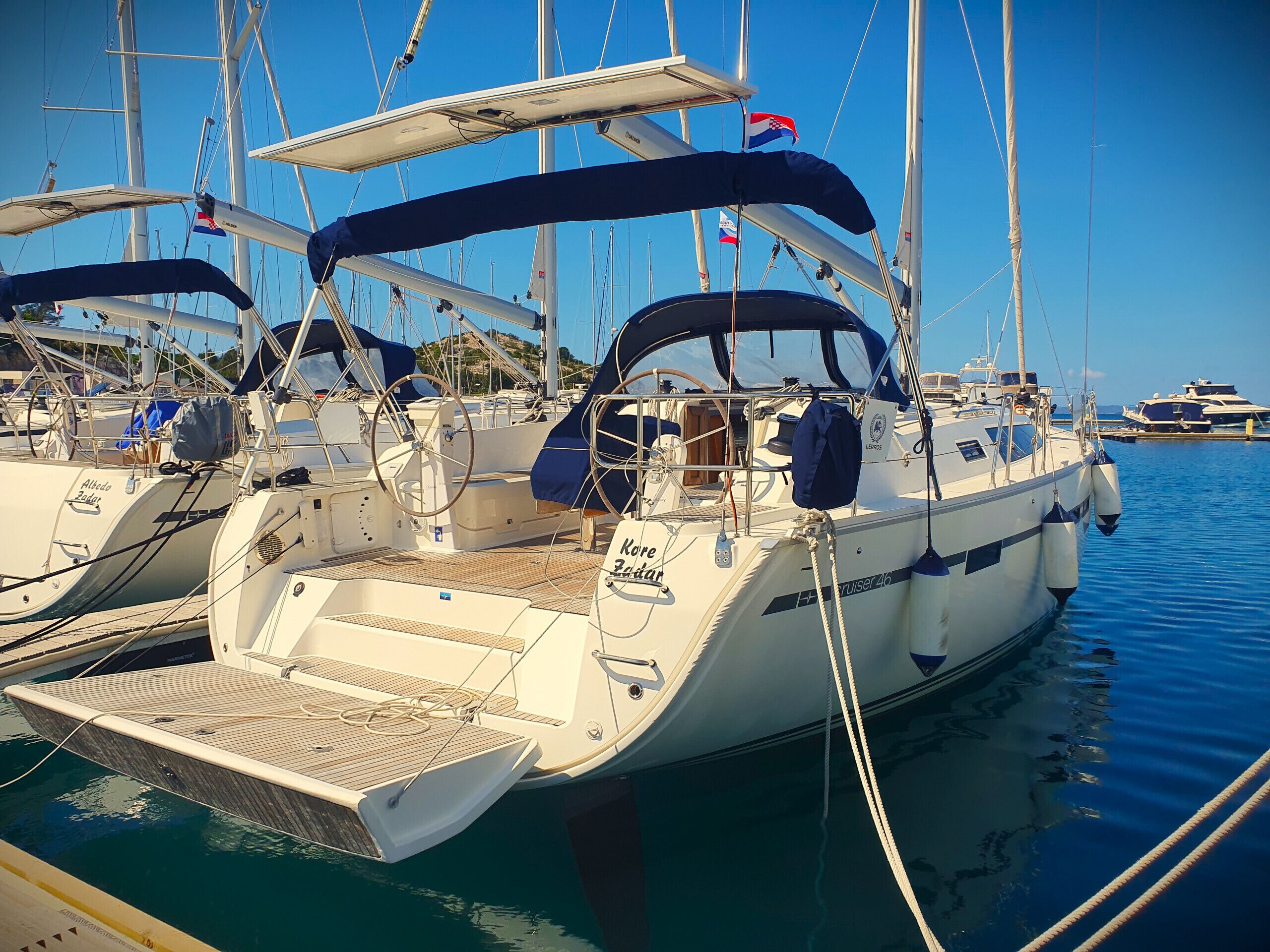 Ein Segelboot liegt in einer Marina festgemacht, mit mehreren Masten und Flaggen, einer Kabine mit Fenstern und einem Deck.