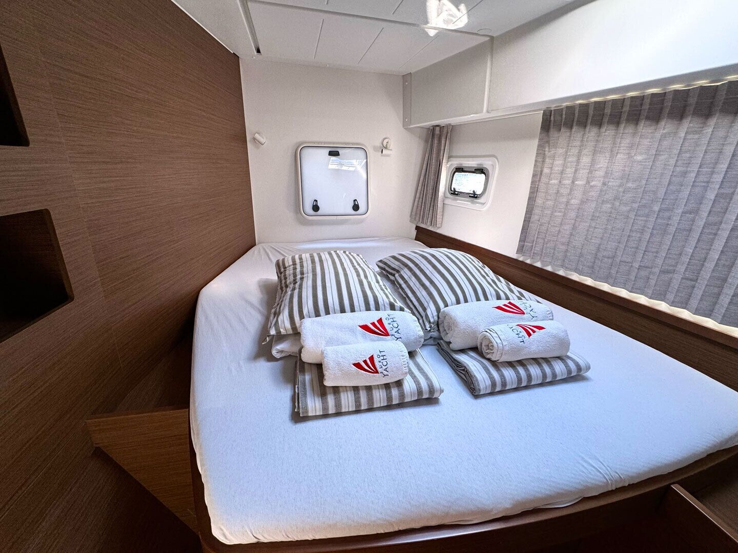 A neatly made bed with striped pillows and towels, a small window with curtains, and a wall-mounted light fixture.