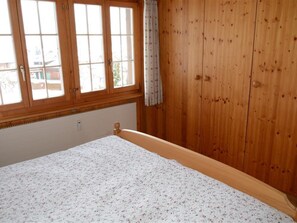 A bedroom with a wooden wardrobe, a bed with a floral bedspread, and a window with curtains. - Résidence Sonnegg (Herrli) Appartement/Fewo, Dusche oder Bad, WC 4-Bettwohnung by Interhome (Zweisimmen)