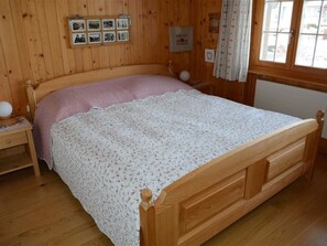 A wooden bedroom with a bed, a nightstand, a wall-mounted photo frame, and a window with curtains. - Résidence Sonnegg (Herrli) Appartement/Fewo, Dusche oder Bad, WC 4-Bettwohnung by Interhome (Zweisimmen)