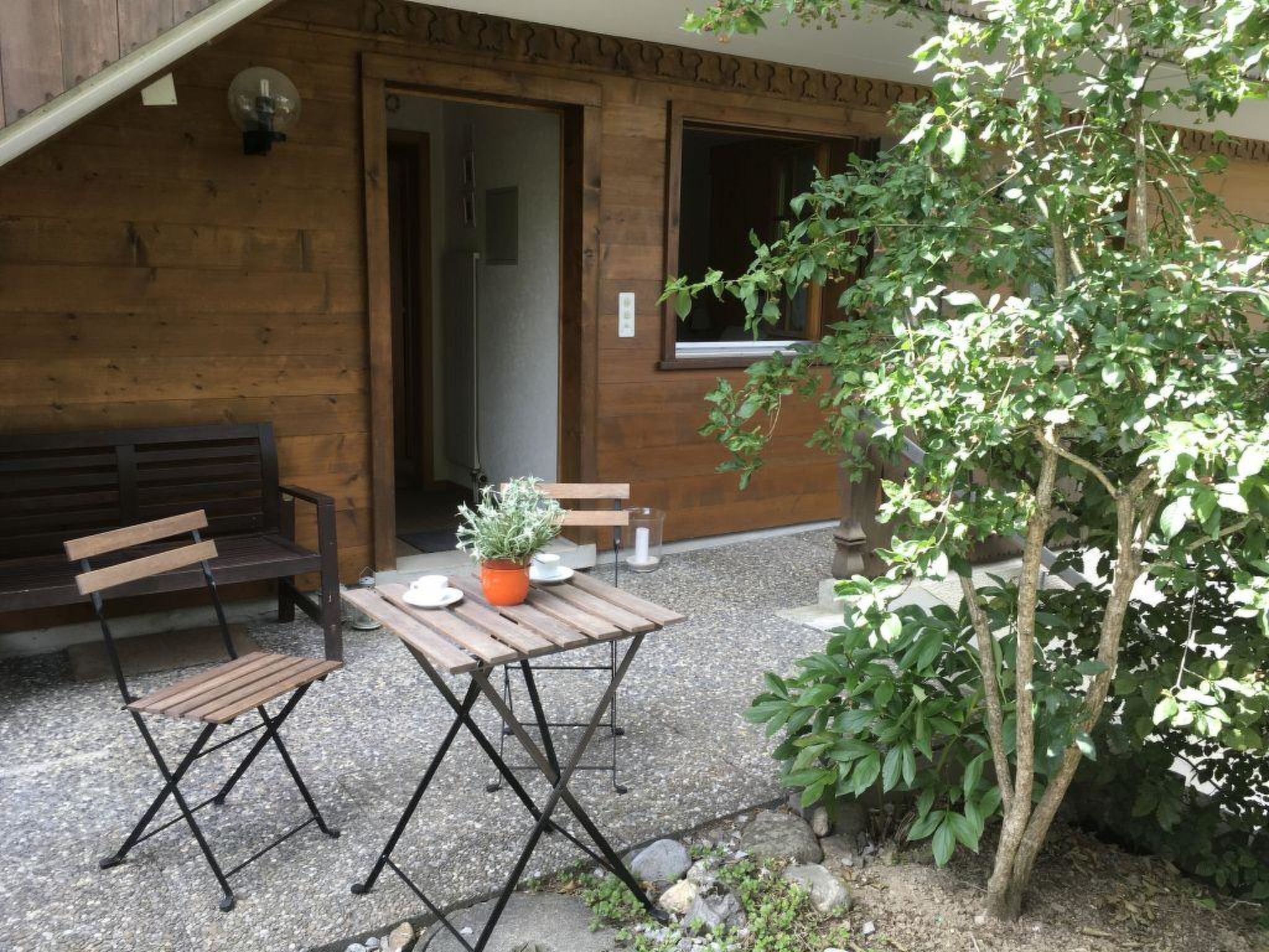 Una pared exterior de madera, un banco, una mesa con una planta en maceta y un árbol.