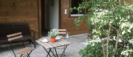 Una pared exterior de madera, un banco, una mesa con una planta en maceta y un árbol.