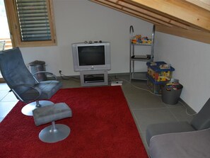 A small living area with a television, a chair, a red rug, and a shelf with items. - Alpha Appartement/Fewo, Dusche oder Bad, WC 4-Bettwohnung by Interhome (Zweisimmen)