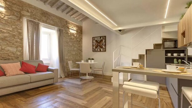 Eine moderne Küche mit einer zentralen Kochinsel, ein Essbereich mit weißem Tisch und Stühlen und eine Wohnbereits mit einer Couch und Kissen.