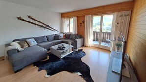 Ett modernt vardagsrum med en grå soffa, ett soffbord och en svartvit matta av kohud. Det finns två fönster med gardiner, ett trägolv och utsikt över omgivningarna.