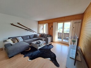 Ein modernes Wohnzimmer mit einem grauen Sofa, einem Couchtisch und einem schwarz-weißen Kuhfellteppich. Zwei Fenster mit Vorhängen, eine Holzdecke und Blick ins Freie.