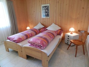 Uma cama de casal com colcha florida, duas mesas de cabeceira com luminárias, uma cadeira de madeira e cabeceira de madeira.