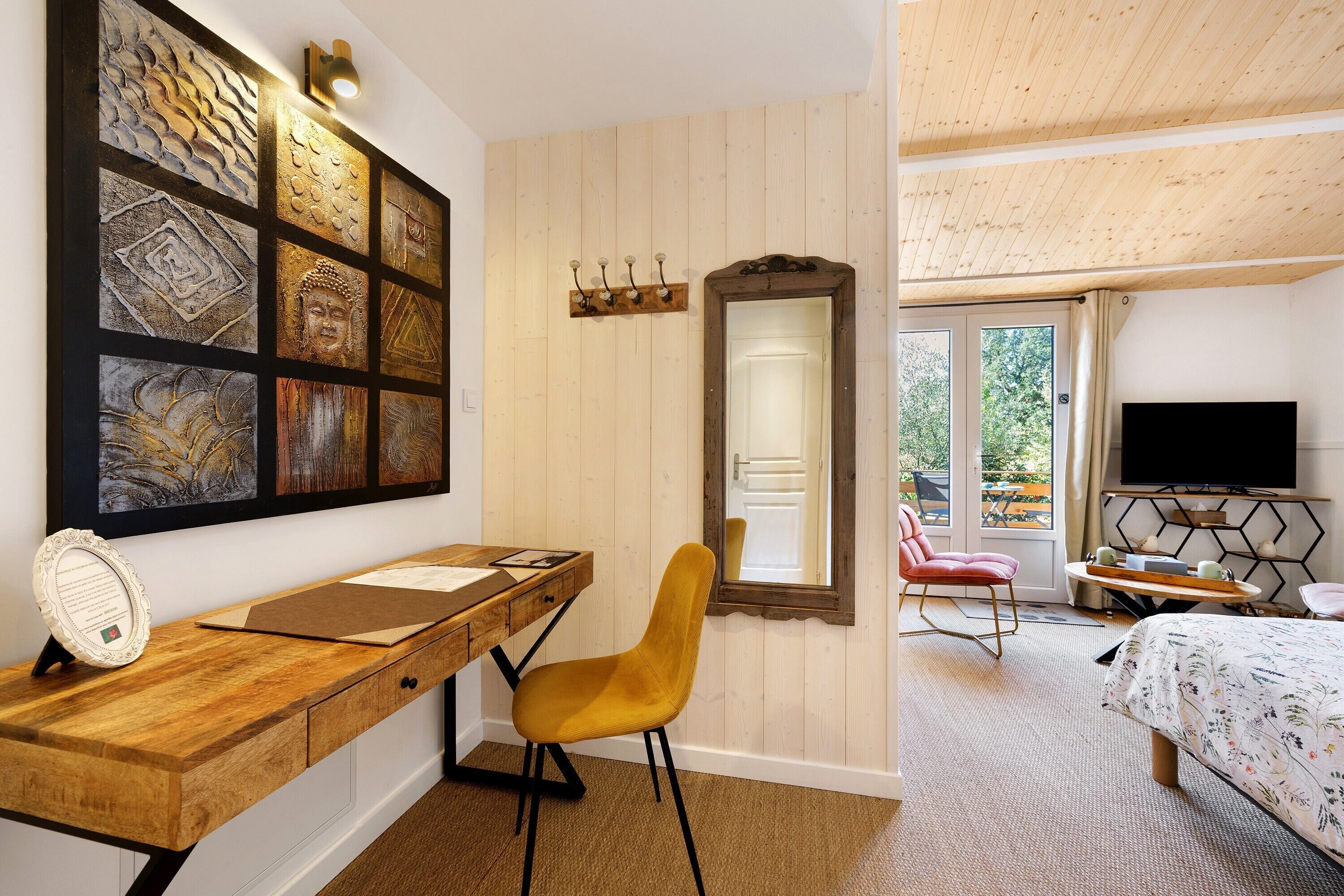 Een kamer met een houten bureau, een stoel, een bed, een televisie en een wand met kunstwerken.