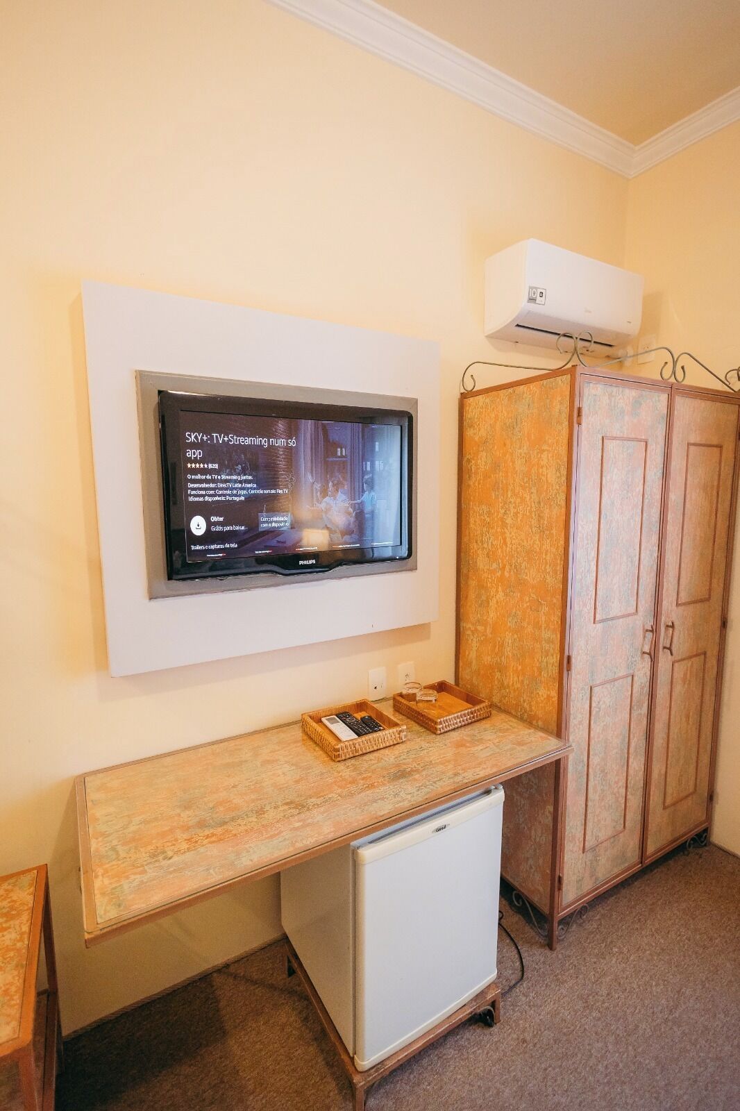 A room with a mounted TV displaying a streaming app interface, a small white refrigerator, a wooden cabinet, and a wooden desk with two wicker baskets.