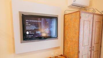 A room with a mounted TV displaying a streaming app interface, a small white refrigerator, a wooden cabinet, and a wooden desk with two wicker baskets.
