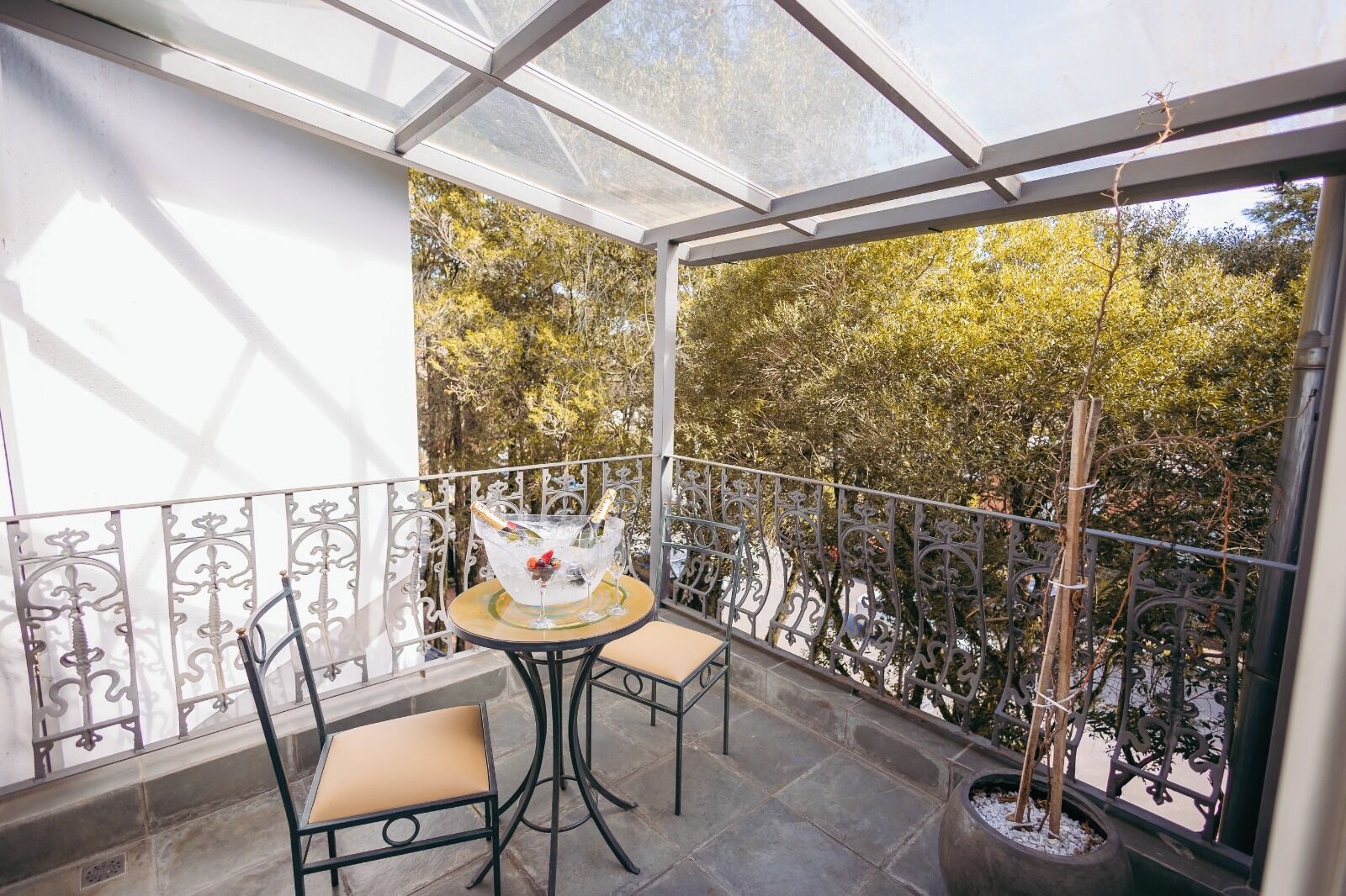 A balcony with a glass enclosure, two chairs, a table with a drink, and a potted plant.