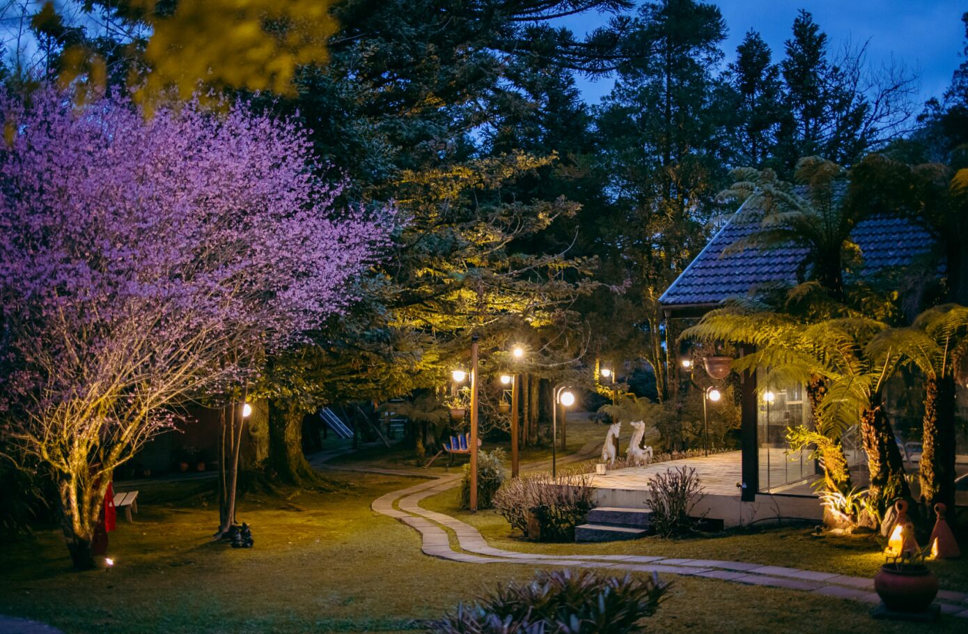 A garden pathway leading to a gazebo with a tiled roof, surrounded by trees and illuminated by street lamps.
