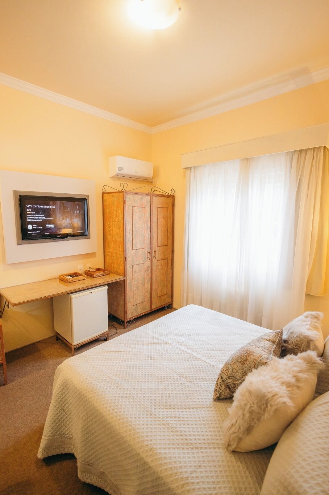 A bedroom with a bed, a desk with a TV, a wardrobe, and a window with curtains.
