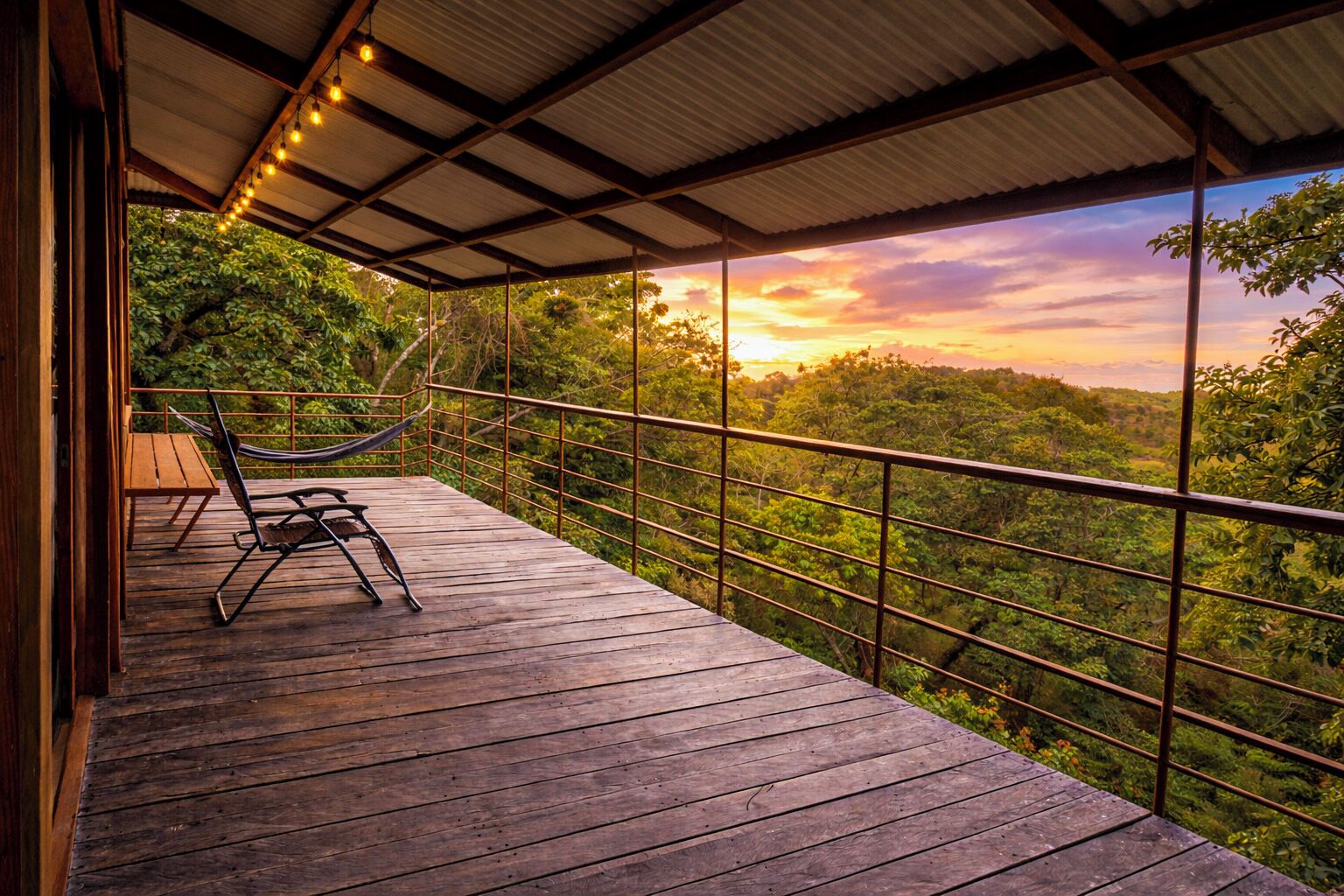 Une terrasse en bois avec un hamac et des chaises, offrant une vue sur un paysage boisé et un magnifique coucher de soleil.