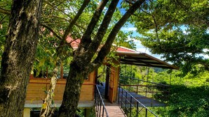 A wooden deck leading to a building with a red roof, surrounded by lush greenery. - El Refugio La Brisa del Diablo – Jungle House (Letrero,)