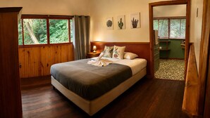 A bedroom with a wooden floor, a bed with a grey and white bedspread, a nightstand with a lamp, and a window with a view of greenery. - El Refugio La Brisa del Diablo – Jungle House (Letrero,)