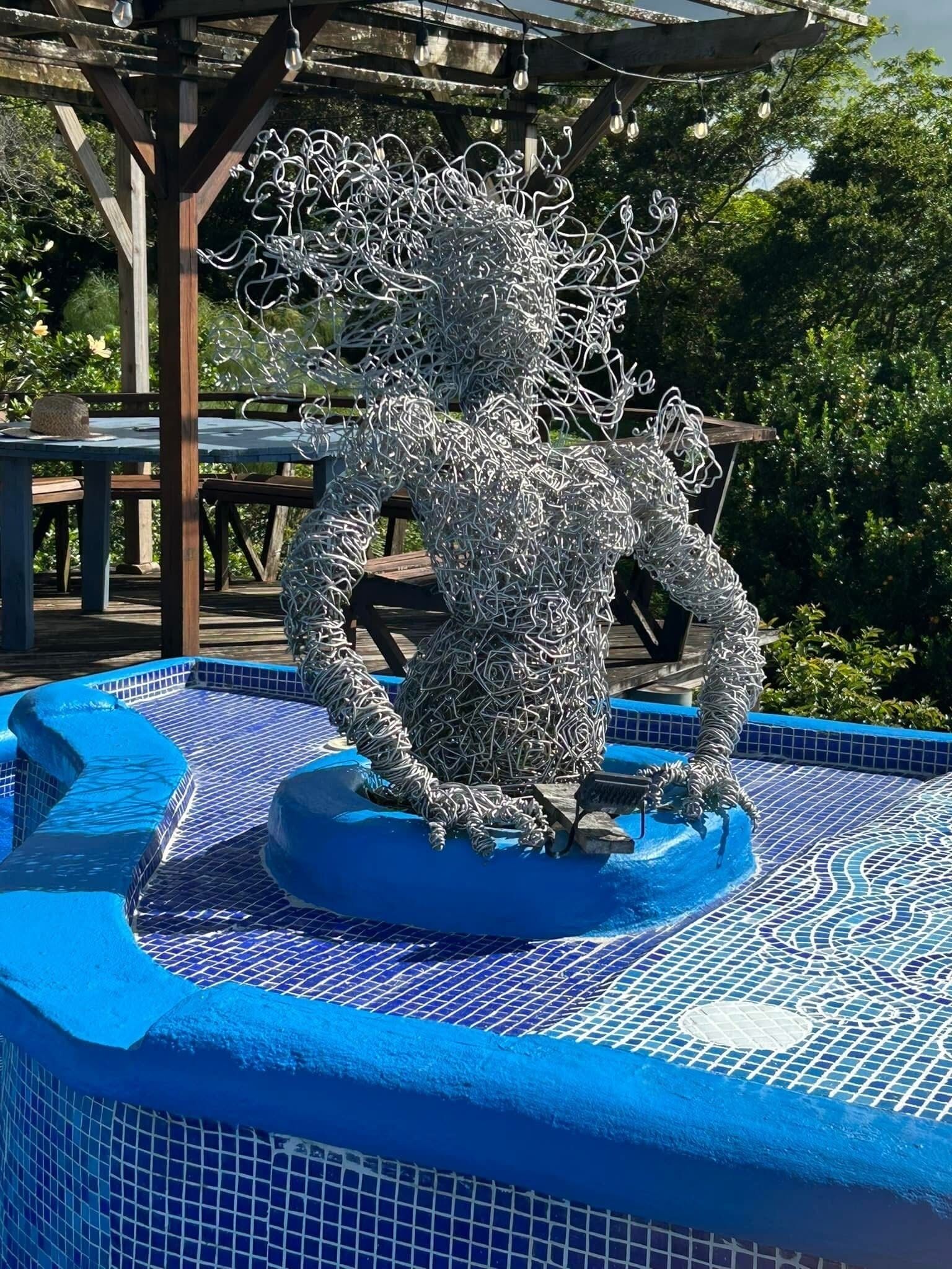 A sculpture of a figure made of intertwined metal wires, positioned on a blue tiled pool with a wooden pergola in the background.
