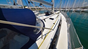Marina con varios veleros amarrados, un barco blanco con una cubierta azul y una dirección web en el mástil.