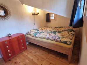 A bedroom with a wooden bed, a dresser, a wall-mounted light, and a window with curtains. - Bachhüsi Appartement/Fewo, Dusche oder Bad, WC 6-Bettwohnung by Interhome (Zweisimmen)
