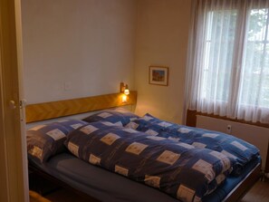 A bedroom with a bed, a bedside lamp, a window with sheer curtains, and a framed picture on the wall.
