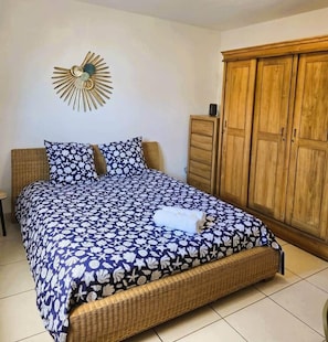 A bed with a floral-patterned bedspread, a decorative wall piece above it, and a wooden wardrobe to the right. - Villa Ö Schandél (Saint-Paul)