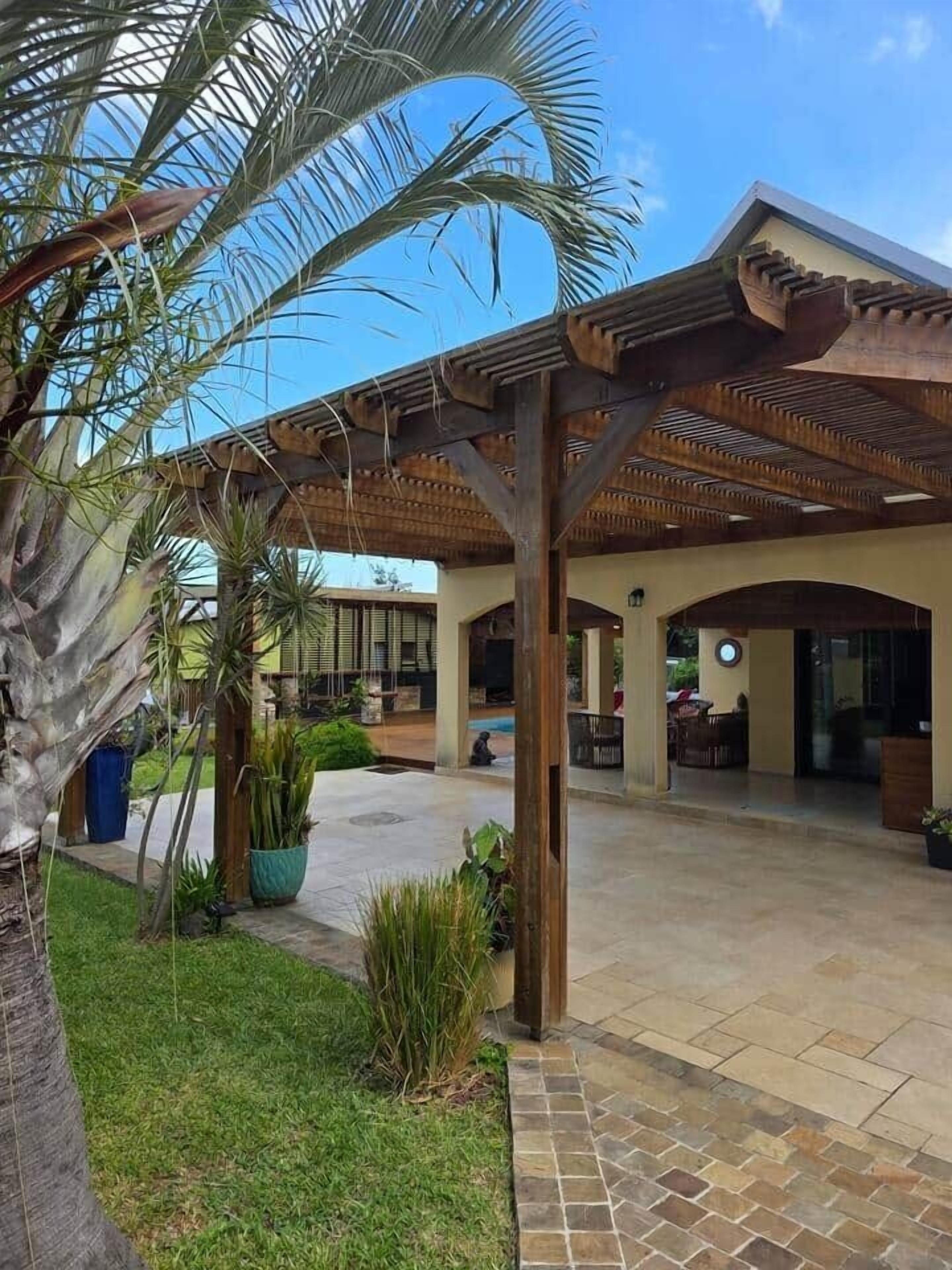 Ein überdachter Außenbereich mit einer Holz-Pergola, Palmen und einem Pool im Hintergrund.