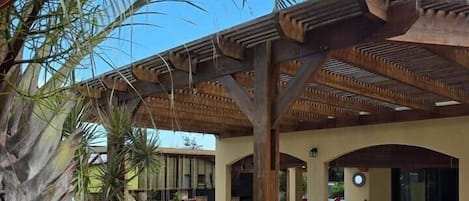 Ein überdachter Außenbereich mit einer Holz-Pergola, Palmen und einem Pool im Hintergrund.