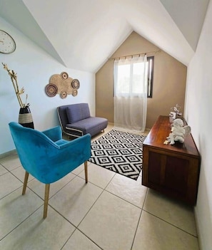 A small, cozy room with a sloped ceiling, a blue chair, a purple sofa, a wooden side table, and a patterned rug. - Villa Ö Schandél (Saint-Paul)
