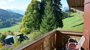 Un balcone in legno con una sedia bianca, affacciato su un paesaggio verde con alberi e montagne in lontananza.