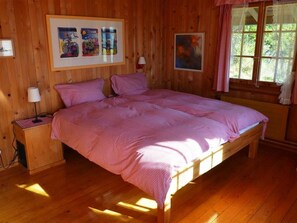 Ein Zimmer mit Holzboden, einem Bett, einem Nachttisch, einer Lampe und einem Fenster mit Vorhängen.