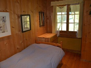 A wooden room with a bed, a small desk, a window with curtains, and a framed picture on the wall.