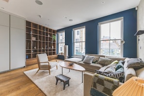 Ein modernes Wohnzimmer mit Sofa, Sessel und Couchtisch, einem Bücherregal und großen Fenstern mit Blick auf die Stadt.