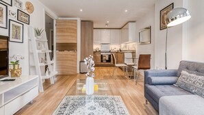 A modern living room with a sofa, a coffee table, and a TV. There is a kitchen in the background with cabinets and a dining area. - Modern Surrey Quays Flat (London)