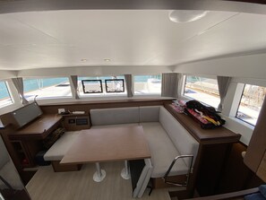 A boat cabin with a table, cushioned seating, a control panel, and windows with curtains.