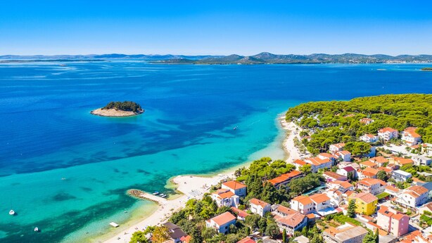 An aerial view of a coastal town with clear blue waters, a small island, and a densely built area with red-roofed buildings.