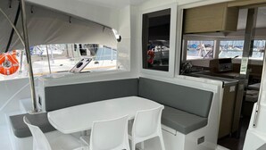 Interno di una barca con tavolo e sedie bianche, una panca grigia e vista sul porto attraverso le finestre.