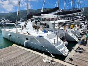 Un puerto deportivo con varios yates amarrados, una cubierta de madera y un paisaje montañoso al fondo.
