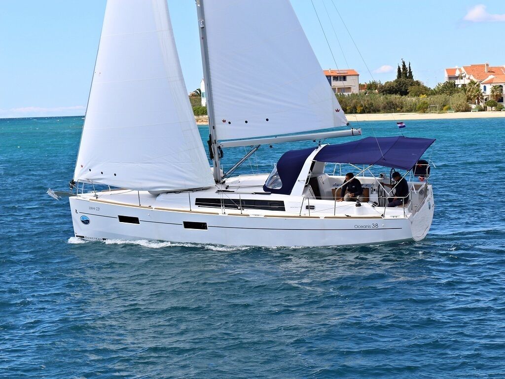 A sailboat with a cabin and a covered deck area, sailing on a body of water with buildings in the background.