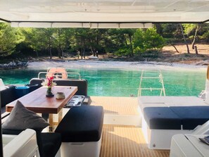 Cubierta de barco con asientos, una mesa y vista a una playa y al agua.