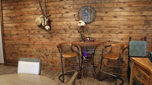 Een rustiek interieur met houten paneelwanden, een ronde houten tafel met twee stoelen, een opgehangen dierenhoofd en een klok aan de muur.