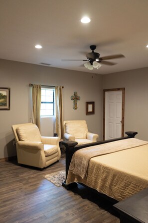 A bedroom with a bed, two chairs, a ceiling fan, a window with curtains, a cross on the wall, and a door.
