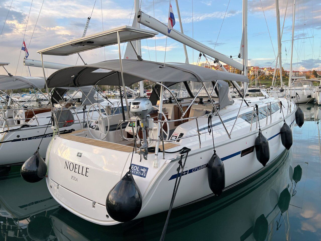 Een witte jacht genaamd 'NOELLE' ligt aangemeerd in een jachthaven, met andere boten, een overdekt dek en een stadsgezicht op de achtergrond.