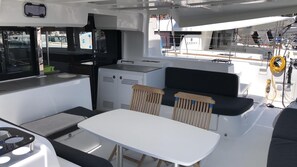 Ein Bootinterior mit einem weißen Tisch, zwei Holzstühlen, einer kleinen Küchenzeile und einer Sitzecke mit einem schwarzen Kissen.