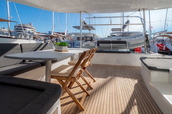 A covered outdoor seating area with tables and chairs, a wooden deck, and a view of other boats in the marina.