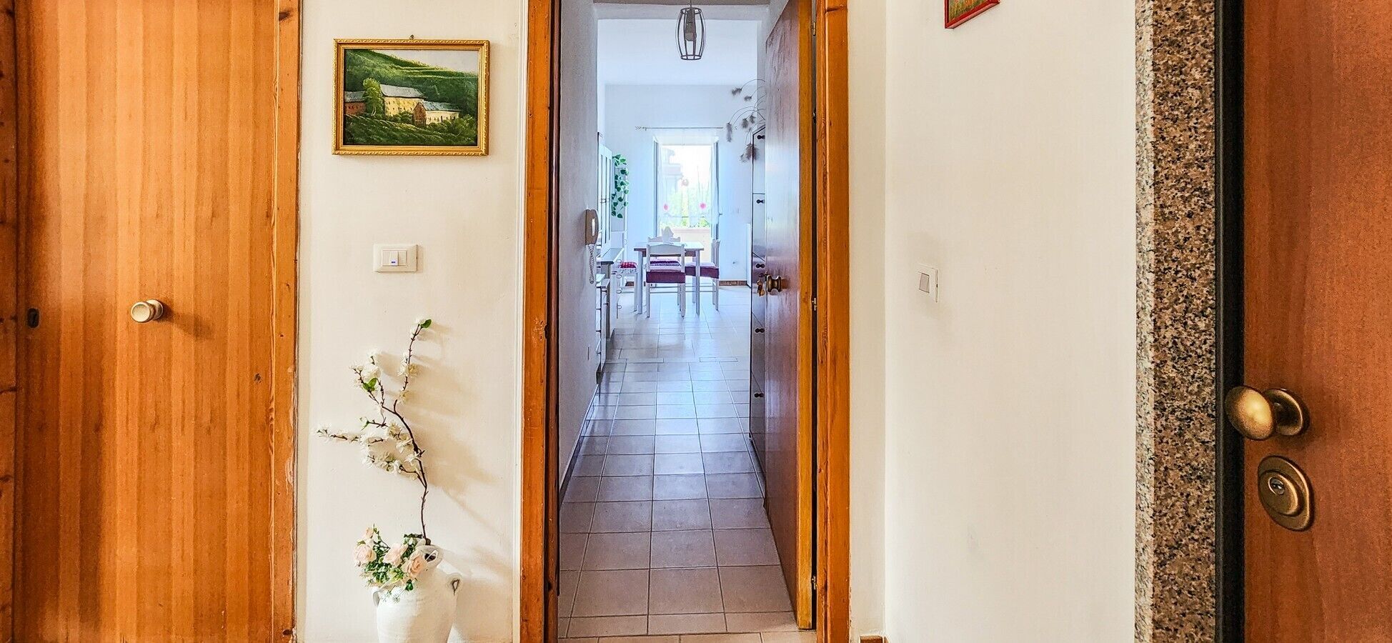A hallway with a wooden door, a framed picture, a small plant, and a tiled floor leading to a dining area.