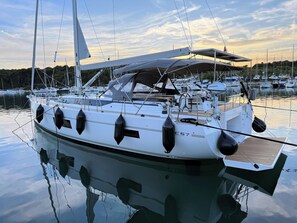 Eine weiße Yacht liegt in einer Marina festgemacht, mit einer überdachten Deckfläche, mehreren schwarzen Fender und einem sichtbaren Motor mit der Aufschrift 'HONDA'.