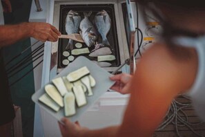 En person som håller ett fat med skivad grönsaker och står bredvid en öppen ugn där det finns fisk och potatis.