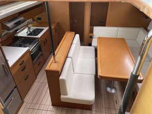 A compact kitchenette with a stove, sink, and cabinets, a wooden dining table with a bench, and a small seating area with a cushioned bench. - Sun-Kissed Sailing Adventure in the Sporades Islands from Volos, Thessaly (Volos)