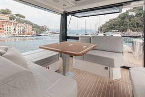 Bootinterieur met een tafel, beklede zitplaatsen en uitzicht op een jachthaven vol boten en gebouwen.
